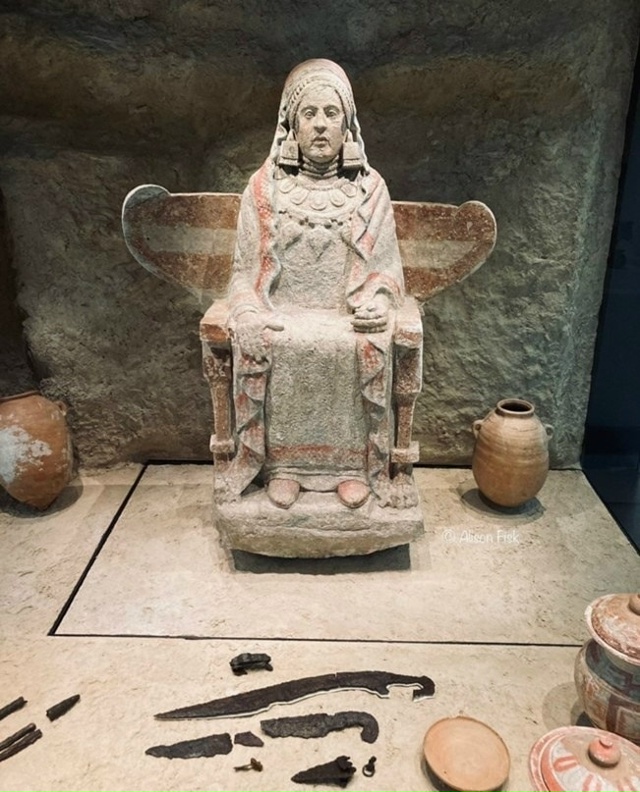 Статуя изображает женщину, сидящую в кресле, причем сб?...